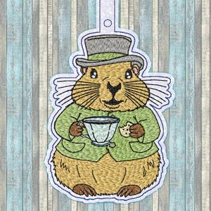 Può includere: Toppa ricamata di un topo marrone e bianco che indossa una giacca verde e un cappello a cilindro, tenendo una tazza da tè e un biscotto.