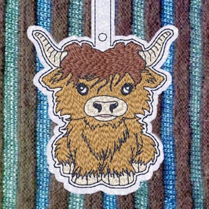 May include: Embroidered design of a brown Highland cow with a white face and long, shaggy hair. The cow is standing with its legs together and its head tilted slightly to the side. The design is perfect for a keychain or ornament.
