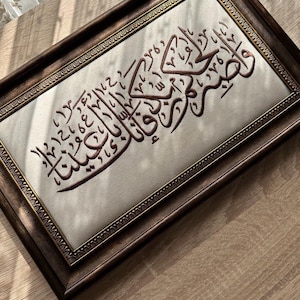 Pode incluir: Uma peça de arte de parede retangular com caligrafia árabe castanho-escura bordada num fundo de tecido creme claro. A obra está emoldurada numa moldura de madeira castanho-escura com uma borda interior dourada ornamentada e texturizada.