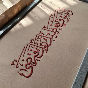 Pode incluir: Um bastidor de bordado retangular contendo tecido bege com caligrafia árabe vermelha costurada no centro. O bordado apresenta uma escrita fluida e complexa com linhas geométricas e pequenos acentos numéricos integrados no design.