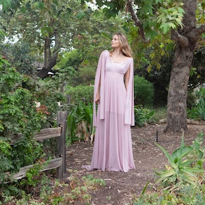 May include: A woman in a long, flowing, light pink dress stands in a garden setting. The dress has a V-neckline and long sleeves. She is looking up and to the right. The background is a lush green garden with a large tree.