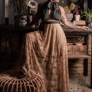 bald woman in olive green and tea cooored lace multiway dress tied in bow around neck standing in front of potter's bench