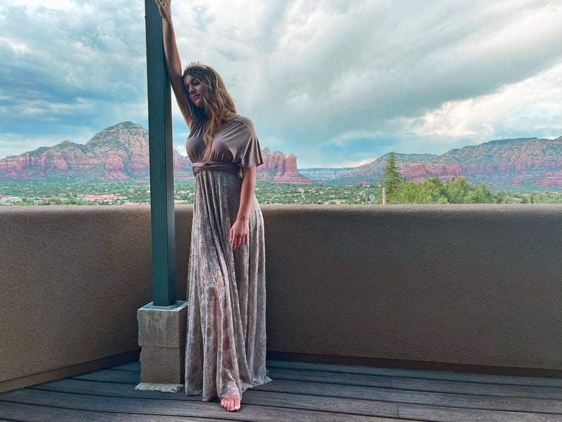 A woman wearing a long, flowing, brown and white dress with a brown belt. She is standing on a wooden deck with a view of red rock mountains in the background.