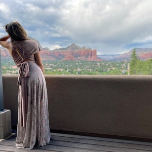 A woman wearing a long, light brown, lace dress with a wrap-style top. The dress has a sash tied at the waist. The woman is standing on a wooden deck overlooking a scenic view of red rock formations and a town in the distance.