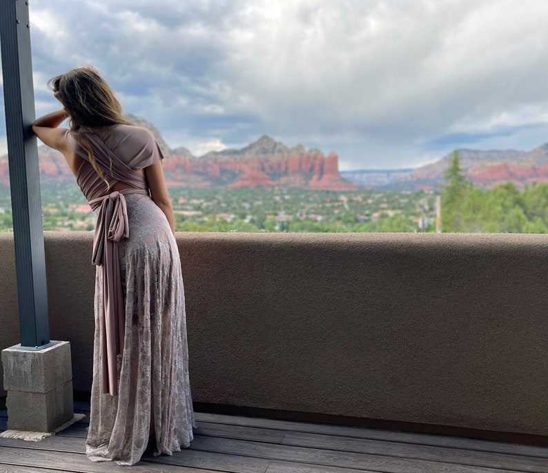 A woman wearing a long, light brown, lace dress with a wrap-style top. The dress has a sash tied at the waist. The woman is standing on a wooden deck overlooking a scenic view of red rock formations and a town in the distance.