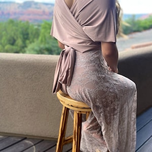 May include: A mauve-colored wrap top with a matching lace-detailed skirt. The top is tied at the waist with a bow. The outfit is worn while seated on a wooden stool. The background features a scenic view of a mountain range.