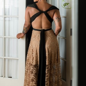 May include: A woman wearing a black and beige maxi dress with a lace overlay. The dress has a low back and a criss-cross strap design. The woman is standing in front of a white door.