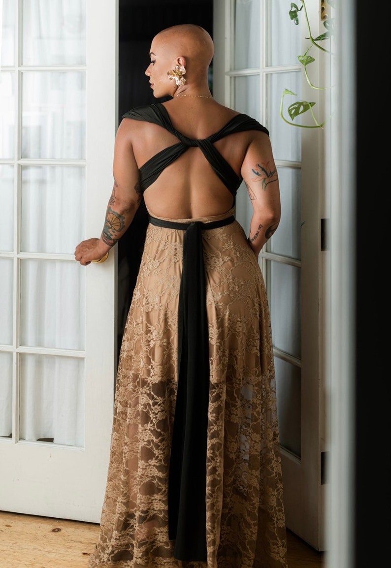 May include: A woman wearing a black and beige maxi dress with a lace overlay. The dress has a low back and a criss-cross strap design. The woman is standing in front of a white door.