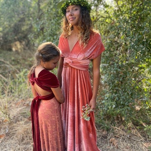 Two girls wearing long, flowing, pink velvet dresses. The girl in the front is wearing a burgundy velvet sash around her waist. The girl in the back is wearing a green leaf crown.