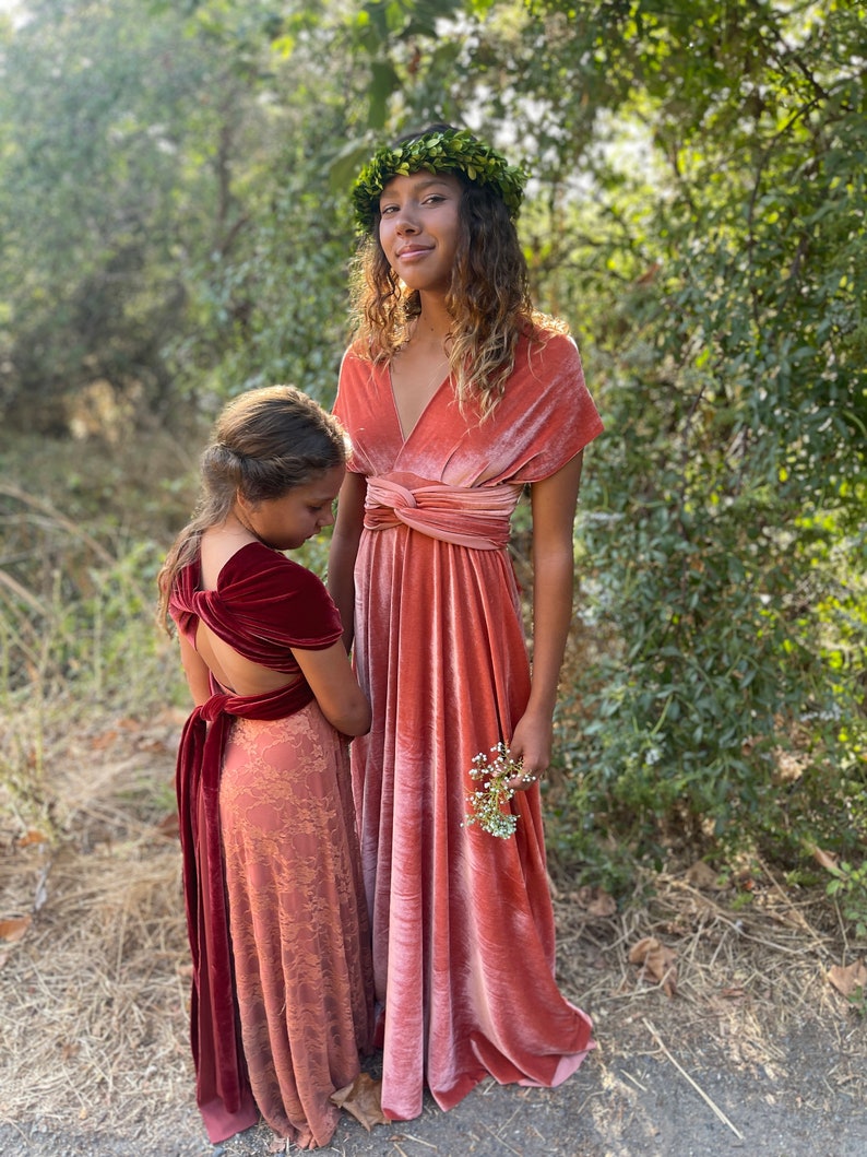 Two girls wearing long, flowing, pink velvet dresses. The girl in the front is wearing a burgundy velvet sash around her waist. The girl in the back is wearing a green leaf crown.