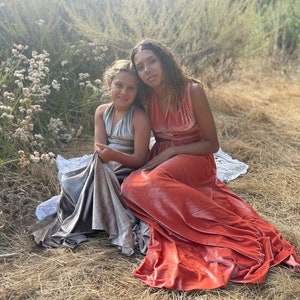 Two girls wearing long, flowing dresses. The girl on the left is wearing a silver dress and the girl on the right is wearing a coral dress.