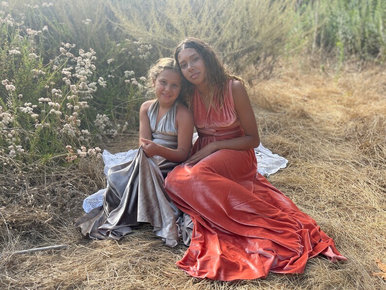 Two girls wearing long, flowing dresses. The girl on the left is wearing a silver dress and the girl on the right is wearing a coral dress.