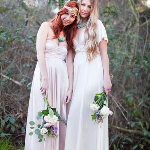 May include: Two individuals in flowing dresses, one blush and one ivory, stand together outdoors. Each holds a bouquet of flowers. They wear decorative headbands and turquoise necklaces, creating a bohemian aesthetic.