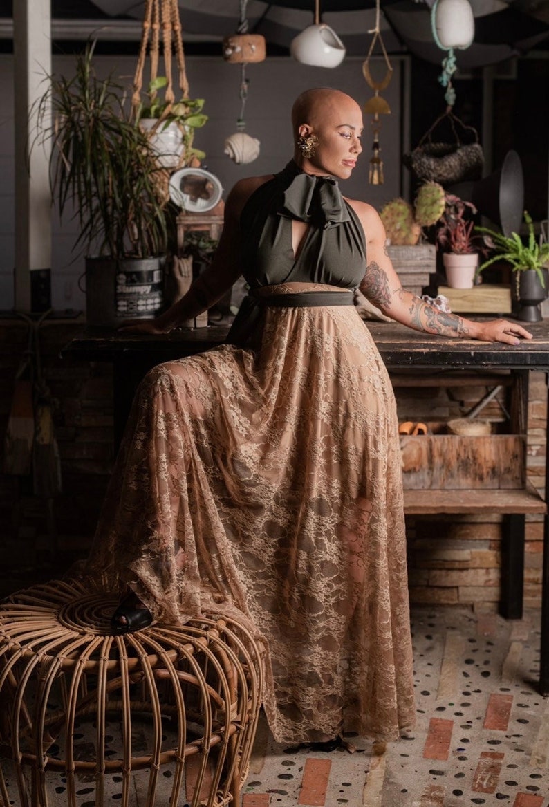 bald woman in olive green and tea cooored lace multiway dress tied in bow around neck standing in front of potter's bench