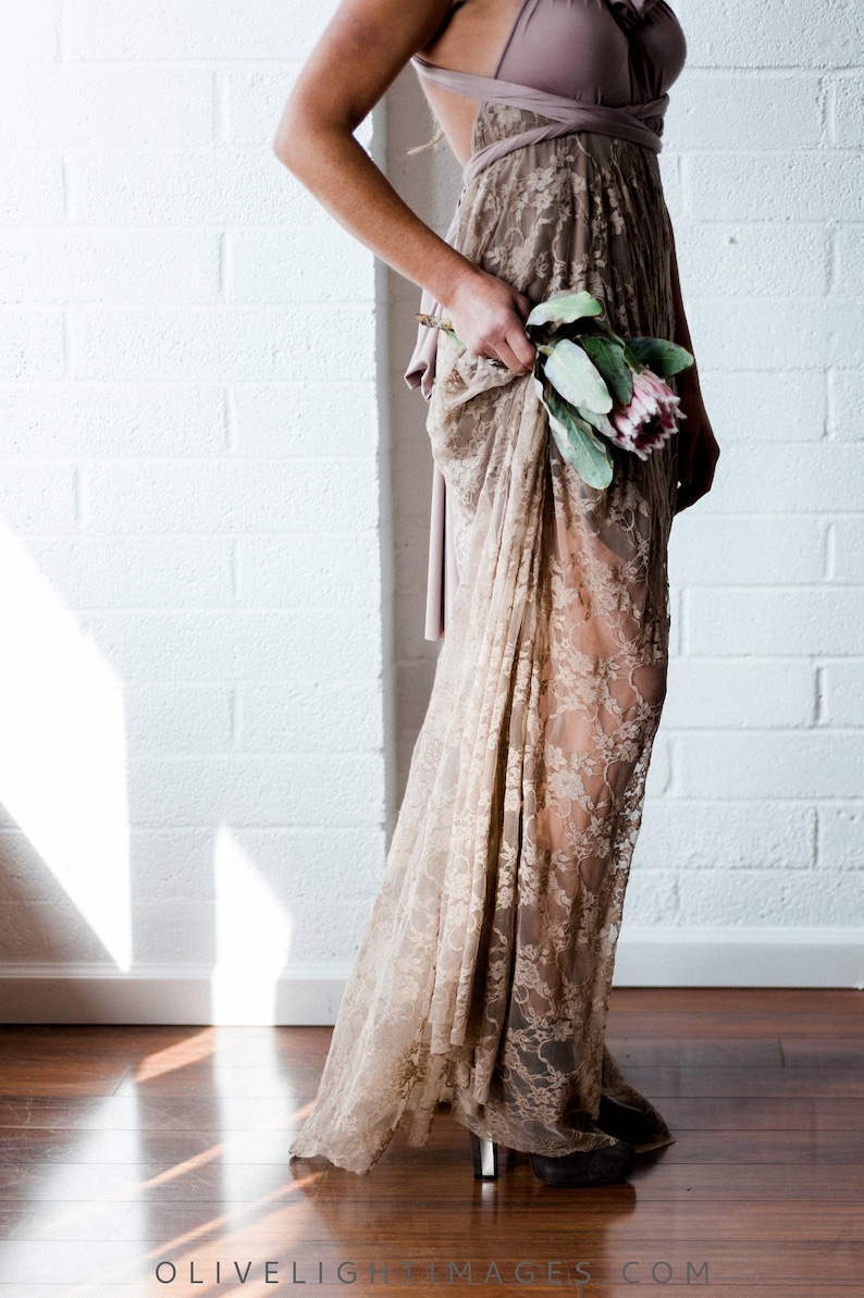 May include: A woman wearing a long, beige lace dress with a brown top. She is holding a bouquet of pink and green flowers.