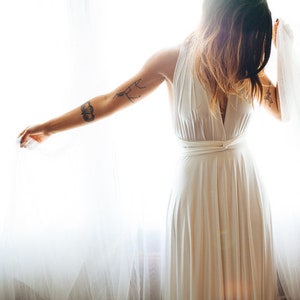 Woman embracing the light coming through the sheers. Dramatic photo of girl with tattoos looking down highlighting the simple beauty of her white Infinity Wrap dress. Worn halter style, looped front at the high waist. Romantic, empowering, sexy.