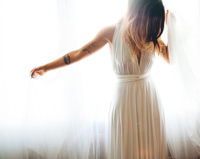 Woman embracing the light coming through the sheers. Dramatic photo of girl with tattoos looking down highlighting the simple beauty of her white Infinity Wrap dress. Worn halter style, looped front at the high waist. Romantic, empowering, sexy.