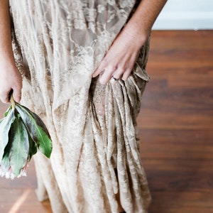 May include: A woman wearing a long, flowing, beige lace dress with a light brown sash. The dress is gathered at the waist and has a flowing skirt. The woman is holding a bouquet of green leaves and pink flowers.