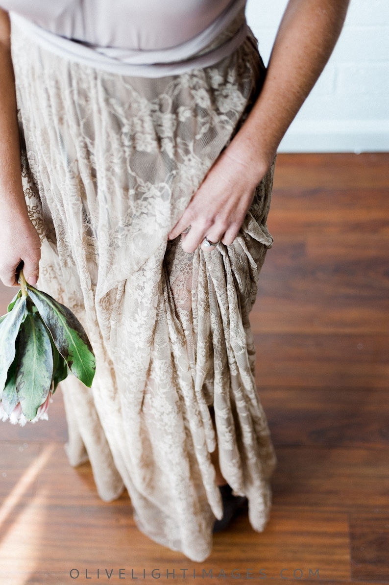 May include: A woman wearing a long, flowing, beige lace dress with a light brown sash. The dress is gathered at the waist and has a flowing skirt. The woman is holding a bouquet of green leaves and pink flowers.