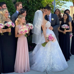 May include: A wedding ceremony with the bride and groom kissing in the centre of the image. The bride is wearing a white lace wedding dress with a veil. The groom is wearing a black suit and a white shirt. The bridal party are standing behind the couple, all wearing formal attire. The bridesmaids are wearing long black and pink dresses and holding bouquets of pink flowers. The groomsmen are wearing black suits and white shirts. The wedding party are standing in front of a brick building with green trees and bushes in the background.