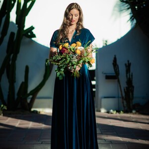 A woman wearing a teal blue velvet dress and holding a bouquet of flowers with yellow, white, and red blooms.