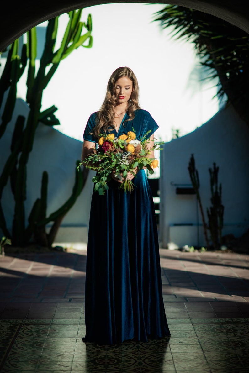 A woman wearing a teal blue velvet dress and holding a bouquet of flowers with yellow, white, and red blooms.