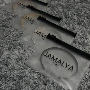 Puede incluir: Varias bolsas de plástico transparente con cremalleras negras, cada una con una fina diadema de metal curvada. El texto "JAMALYA PARIS" está impreso en las bolsas. Las diademas son plateadas y doradas.
