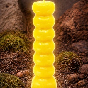 May include: A tall, yellow pillar candle with a unique, stacked design. The candle has a smooth, glossy finish and a single wick at the top. The candle is set against a natural background of moss, rocks, and a pine cone.