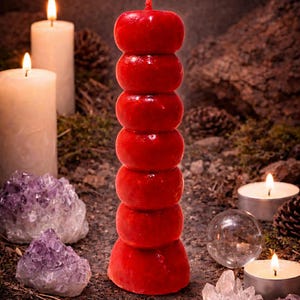 May include: A tall, red candle with a stacked design, resembling a tower, stands amidst lit candles, crystals, and pine cones. The candle's vibrant color and unique shape create a striking visual contrast against the natural elements.