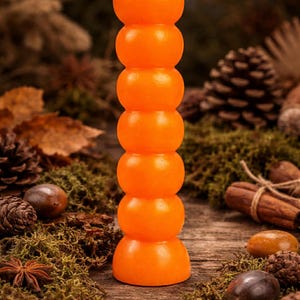 May include: An orange pillar candle with a stacked, rounded design. The candle has a white wick and is surrounded by natural elements like pine cones, moss, and cinnamon sticks. The candle is approximately 6 inches tall.