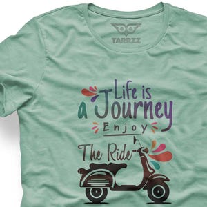 Könnte beinhalten: Hellgrünes T-Shirt mit dem Text "Life is a Journey, Enjoy The Ride" und einer braunen Rollergrafik. Das Design enthält farbenfrohe Akzente und ist auf einem weichen, lässigen T-Shirt gedruckt.