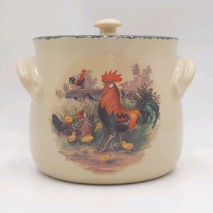 May include: A cream-colored ceramic canister with a lid and two side handles. The front features a colorful illustration of a large rooster, a hen, and several small yellow chicks in a grassy field with trees in the background.