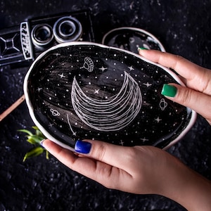 Puede incluir: Un plato de cerámica negro con borde blanco, con un diseño celestial dibujado a mano de una luna creciente, estrellas y planetas. El plato está sostenido por manos con uñas pintadas. Una cámara vintage está en el fondo.