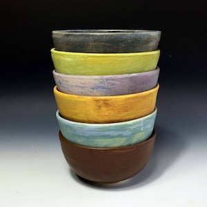 May include: A stack of six ceramic bowls in various colors, including brown, yellow, blue, purple, green, and gray. The bowls are nested inside each other, with the largest bowl at the bottom and the smallest bowl at the top.