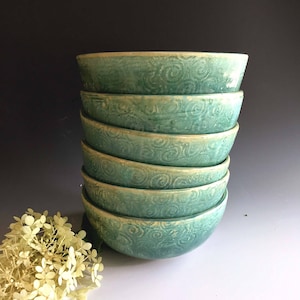 May include: A stack of seven teal ceramic bowls with a swirling pattern. The bowls are stacked on a white surface.