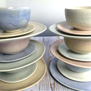 Puede incluir: Apilados de cuencos y platos de cer&aacute;mica en colores pastel. Los cuencos son redondos con un borde ligeramente ensanchado, y los platos son planos con un dise&ntilde;o sencillo. Los colores incluyen azul claro, rosa y beige. La vajilla est&aacute; dispuesta sobre una superficie de madera clara.