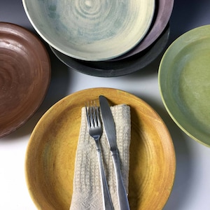May include: A set of five ceramic plates in various colors, including brown, green, yellow, and blue. The plates are stacked on top of each other, with a fork and knife placed on top of the yellow plate.