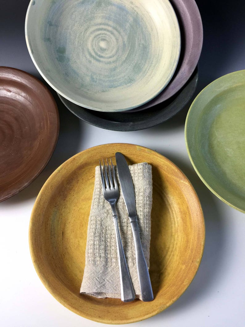 May include: A set of five ceramic plates in various colors, including brown, green, yellow, and blue. The plates are stacked on top of each other, with a fork and knife placed on top of the yellow plate.
