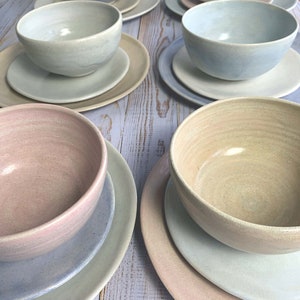 Puede incluir: Colecci&oacute;n de cuencos y platos de cer&aacute;mica hechos a mano en varios colores pastel, incluyendo azul, rosa y beige. Los cuencos se asientan sobre platos a juego, creando un conjunto de vajilla cohesivo y elegante. La cer&aacute;mica tiene un acabado r&uacute;stico y texturizado.