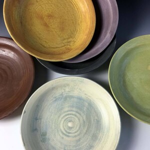 May include: Four ceramic bowls in different colors and patterns. The bowls are stacked on top of each other. The top bowl is yellow with brown streaks, the second bowl is purple, the third bowl is black, and the bottom bowl is green.