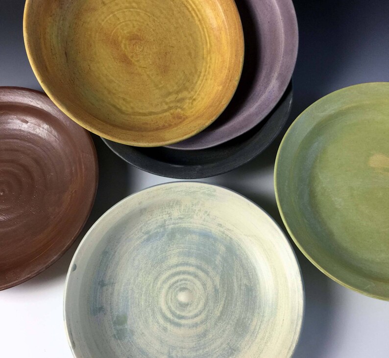 May include: Four ceramic bowls in different colors and patterns. The bowls are stacked on top of each other. The top bowl is yellow with brown streaks, the second bowl is purple, the third bowl is black, and the bottom bowl is green.