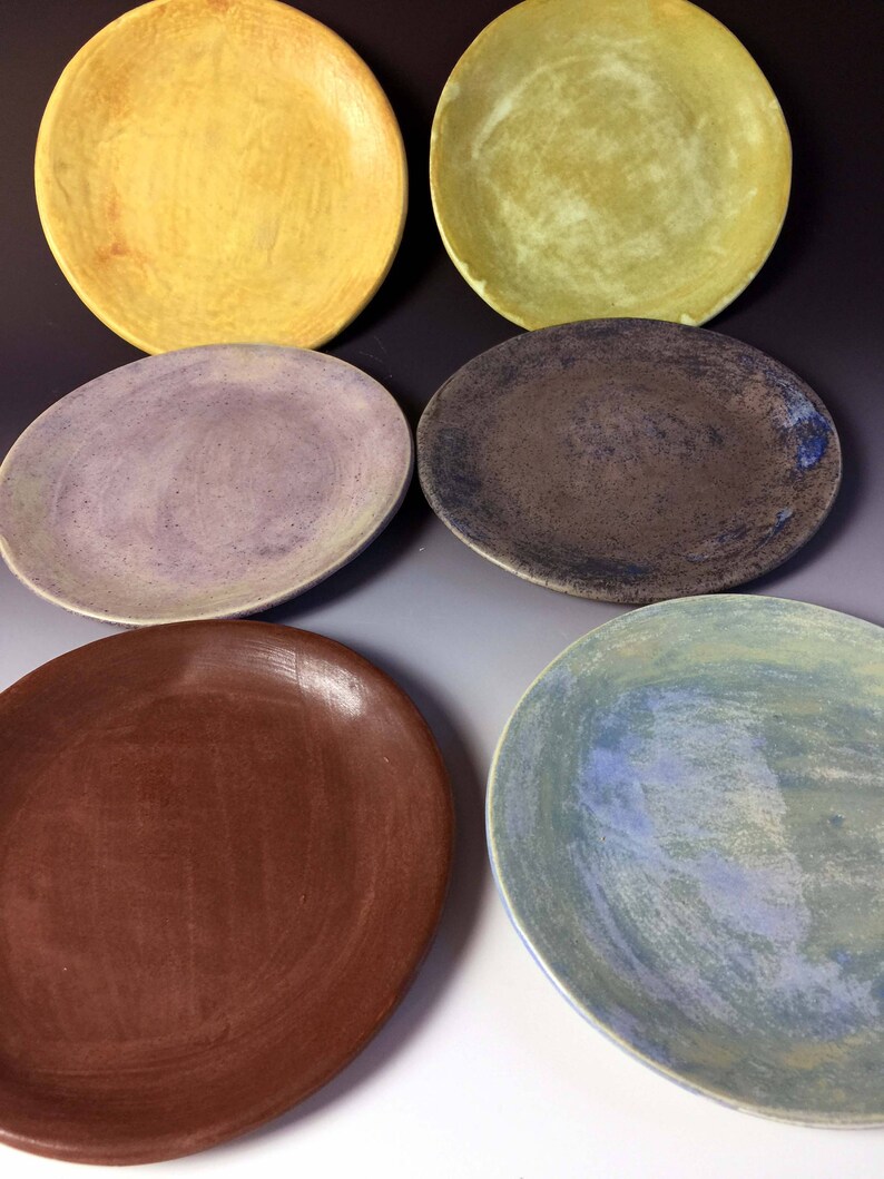 May include: Six ceramic plates in various colors and patterns. The plates are round and have a smooth, glazed finish. The colors include yellow, green, purple, brown, and blue.