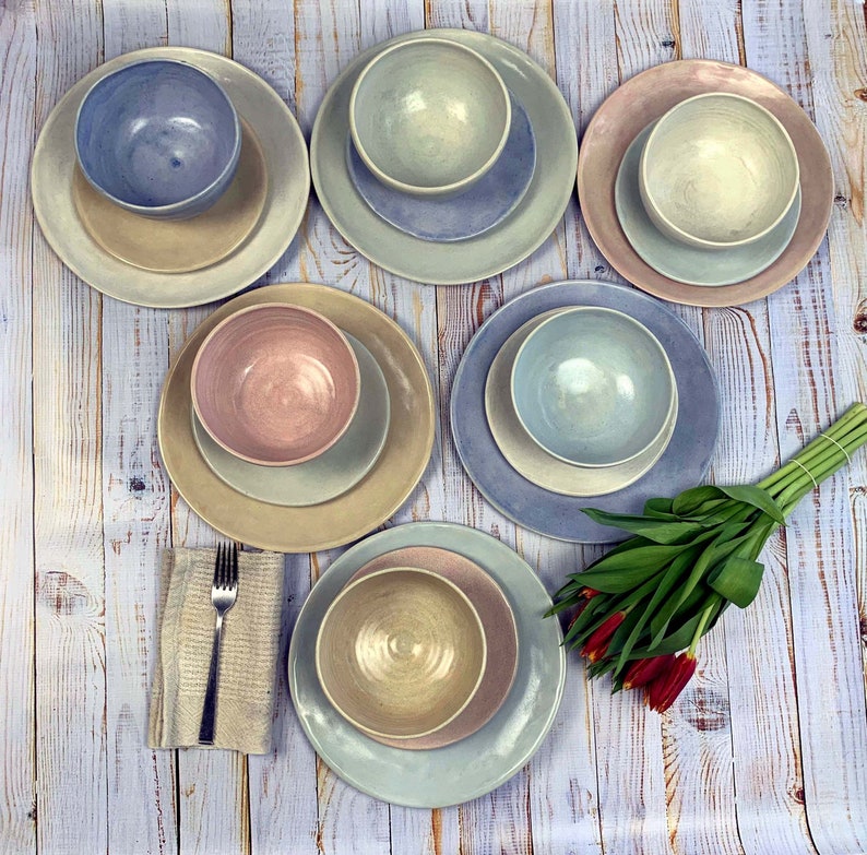 Puede incluir: Conjunto de vajilla de cer&aacute;mica en varios colores pastel, incluyendo azul, rosa y beige. Cada juego incluye un plato grande, un plato m&aacute;s peque&ntilde;o y un cuenco. Un tenedor y una servilleta est&aacute;n a la izquierda, y un ramo de tulipanes rojos a la derecha.