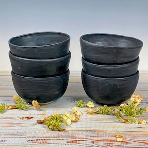 May include: Six black ceramic bowls stacked in pairs. The bowls have a textured surface and a slightly asymmetrical shape. The bowls are arranged on a light-colored wooden surface with dried flowers and greenery.