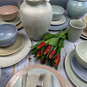 Puede incluir: Colecci&oacute;n de vajilla de cer&aacute;mica artesanal en colores pastel suaves. El conjunto incluye platos, cuencos, una jarra y una taza. Un ramo de tulipanes rojos a&ntilde;ade un toque de color. La cer&aacute;mica tiene un acabado r&uacute;stico y texturizado.