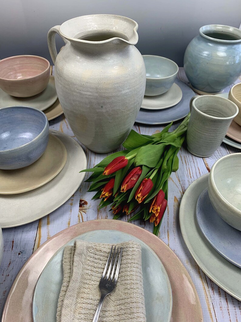 Puede incluir: Colecci&oacute;n de vajilla de cer&aacute;mica artesanal en colores pastel suaves. El conjunto incluye platos, cuencos, una jarra y una taza. Un ramo de tulipanes rojos a&ntilde;ade un toque de color. La cer&aacute;mica tiene un acabado r&uacute;stico y texturizado.