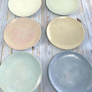 Puede incluir: Seis platos redondos de cer&aacute;mica en varios colores pastel: crema, verde p&aacute;lido, rosa, beige, azul claro y b&iacute;garo. Los platos est&aacute;n colocados sobre una superficie de madera clara.