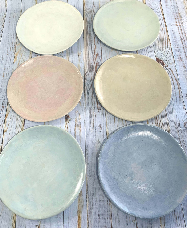 Puede incluir: Seis platos redondos de cer&aacute;mica en varios colores pastel: crema, verde p&aacute;lido, rosa, beige, azul claro y b&iacute;garo. Los platos est&aacute;n colocados sobre una superficie de madera clara.