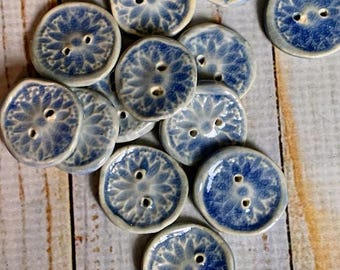 May include: A collection of handmade ceramic buttons. Each button is round with two holes and a blue floral design on a white background. The buttons are scattered on a light wood surface.