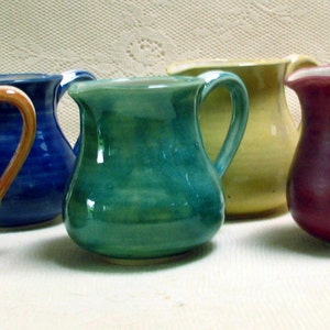 May include: Six small ceramic pitchers in various colors, including orange, blue, green, yellow, and red. The pitchers are all similar in size and shape, with a rounded body and a small spout.